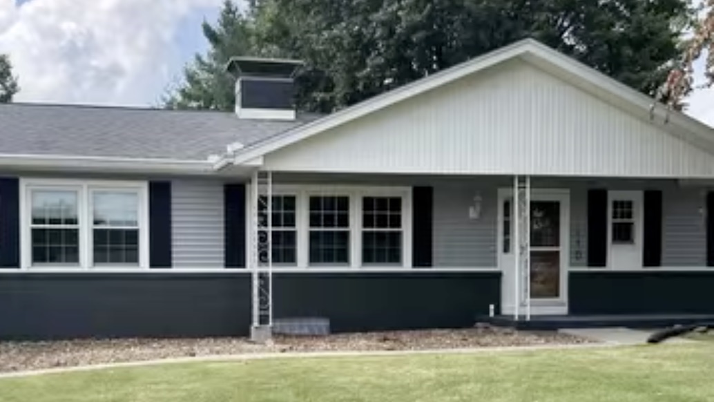 The same house after renovation: the brick base painted dark charcoal, grey siding above it, and black shutters at the windows.