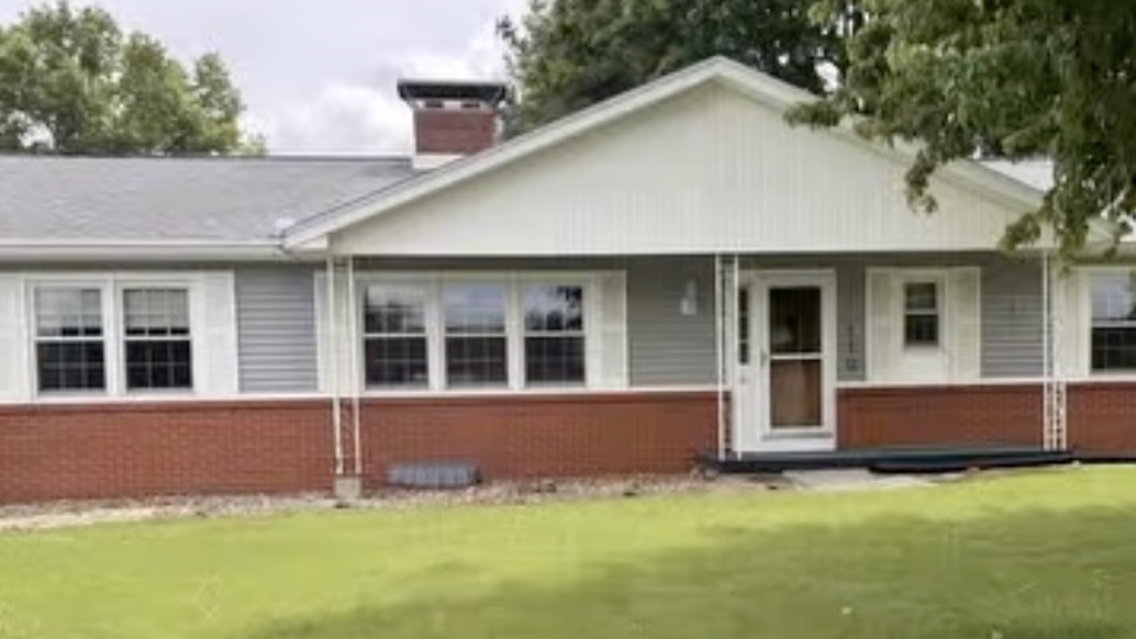 The house before renovation: a single-storey ranch with pale siding, a red brick lower wall and white window and door trim.