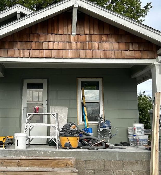 After, with work still under way: the same porch repainted sage green with new cedar shingles in the gable and a new window, and a stepladder, buckets, a shop vacuum, a spirit level and lengths of timber still standing on the porch.
