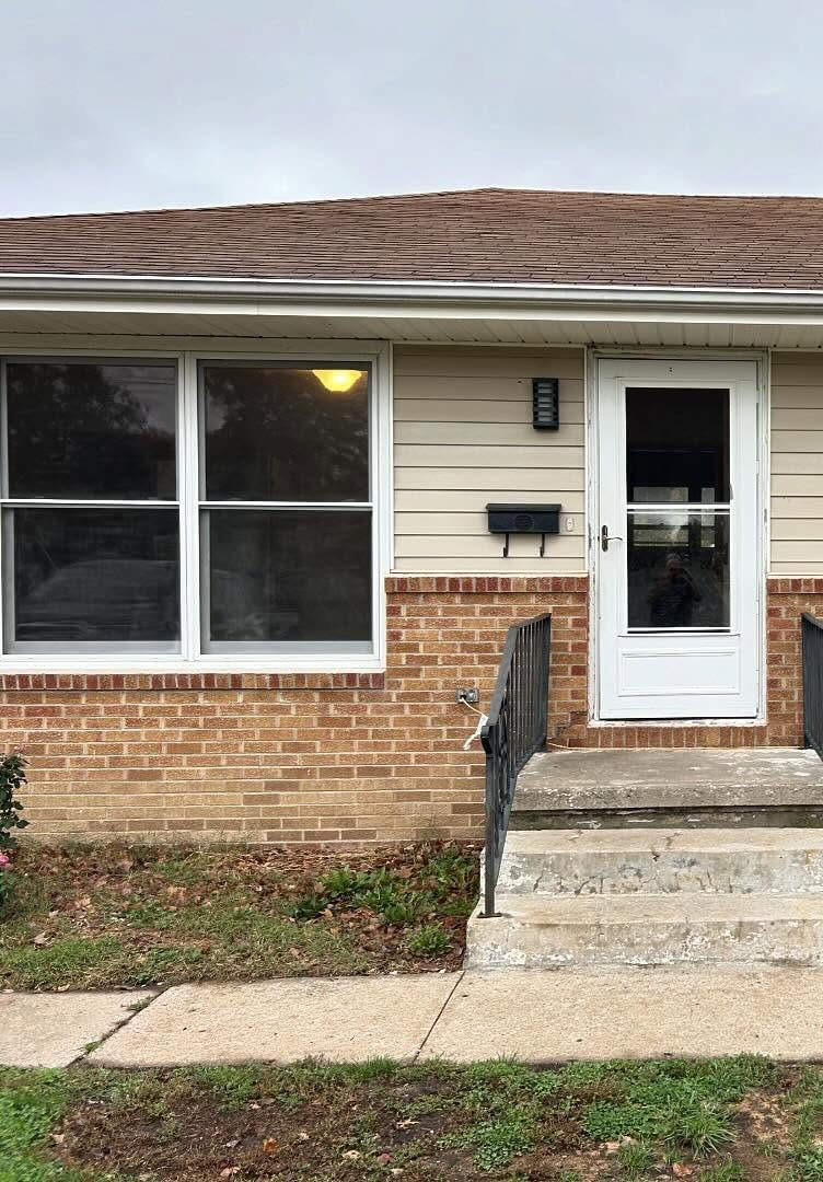 1430 S. English: a red brick lower wall under cream lap siding, a large double window to the left, a white storm door with a wall-mounted mailbox beside it, and concrete steps with a black iron rail.