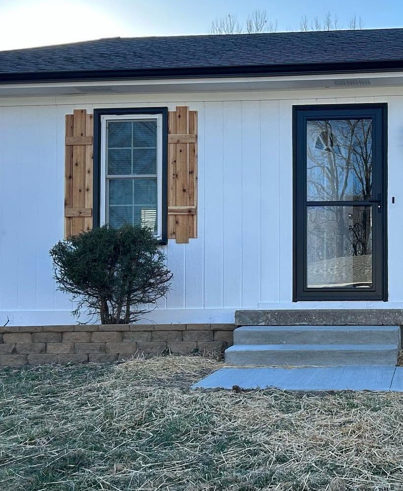 1711 Colby from the front: white board-and-batten siding, a black-trimmed window with rough cedar shutters, a black storm door, a low stone retaining wall and a new concrete walk.