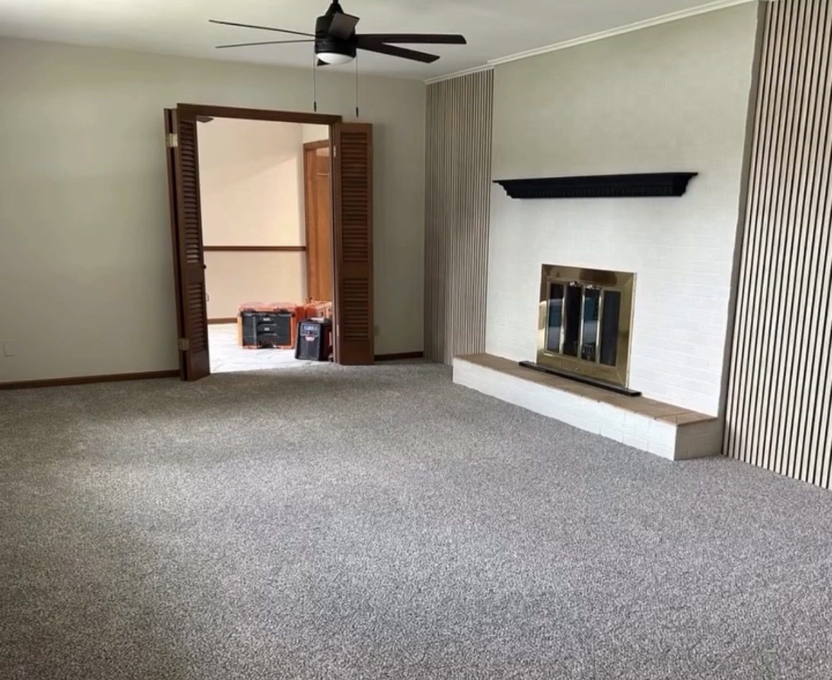 A room mid-renovation: new pale grey carpet, a white-painted brick fireplace with a brass-framed insert and a dark mantel, slatted wood wall panelling, a ceiling fan, and tool cases stacked in the room beyond the louvred doors.