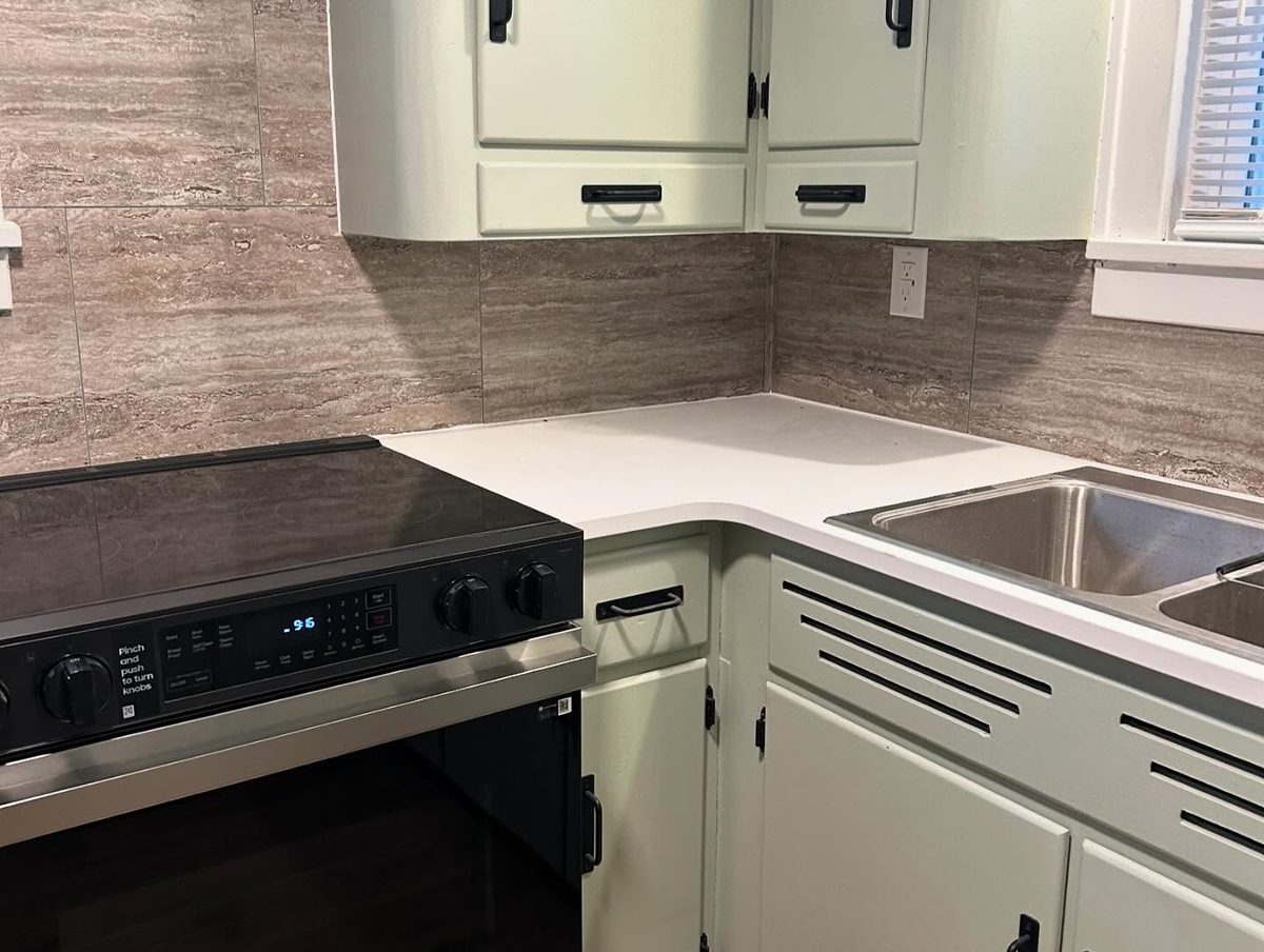 The kitchen at 677 Clay: pale green painted cabinets, a black glass-top electric range, white worktops and a stone-look tiled backsplash beside a stainless steel sink.