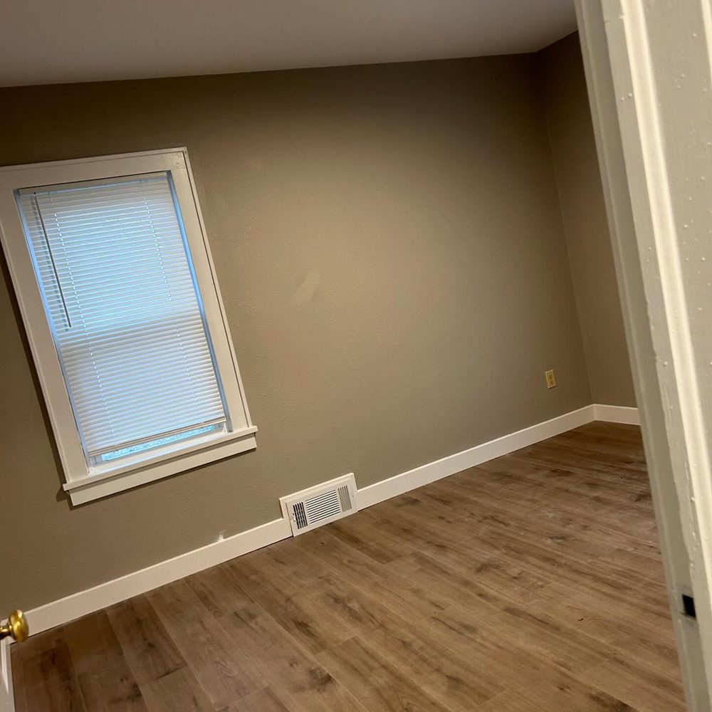 A room at 677 Clay: bare tan walls, white painted skirting and window trim, a blind at the single window, a floor vent, and wood-look plank flooring.