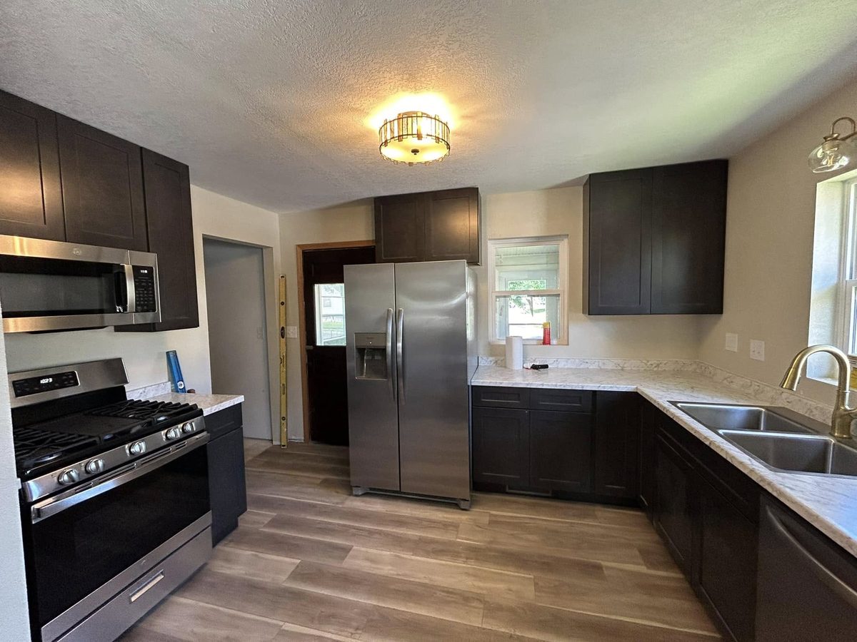 The kitchen of the two-bedroom house: dark brown shaker cabinets, stainless steel range, microwave and side-by-side refrigerator, pale marbled worktops and wood-look plank flooring.